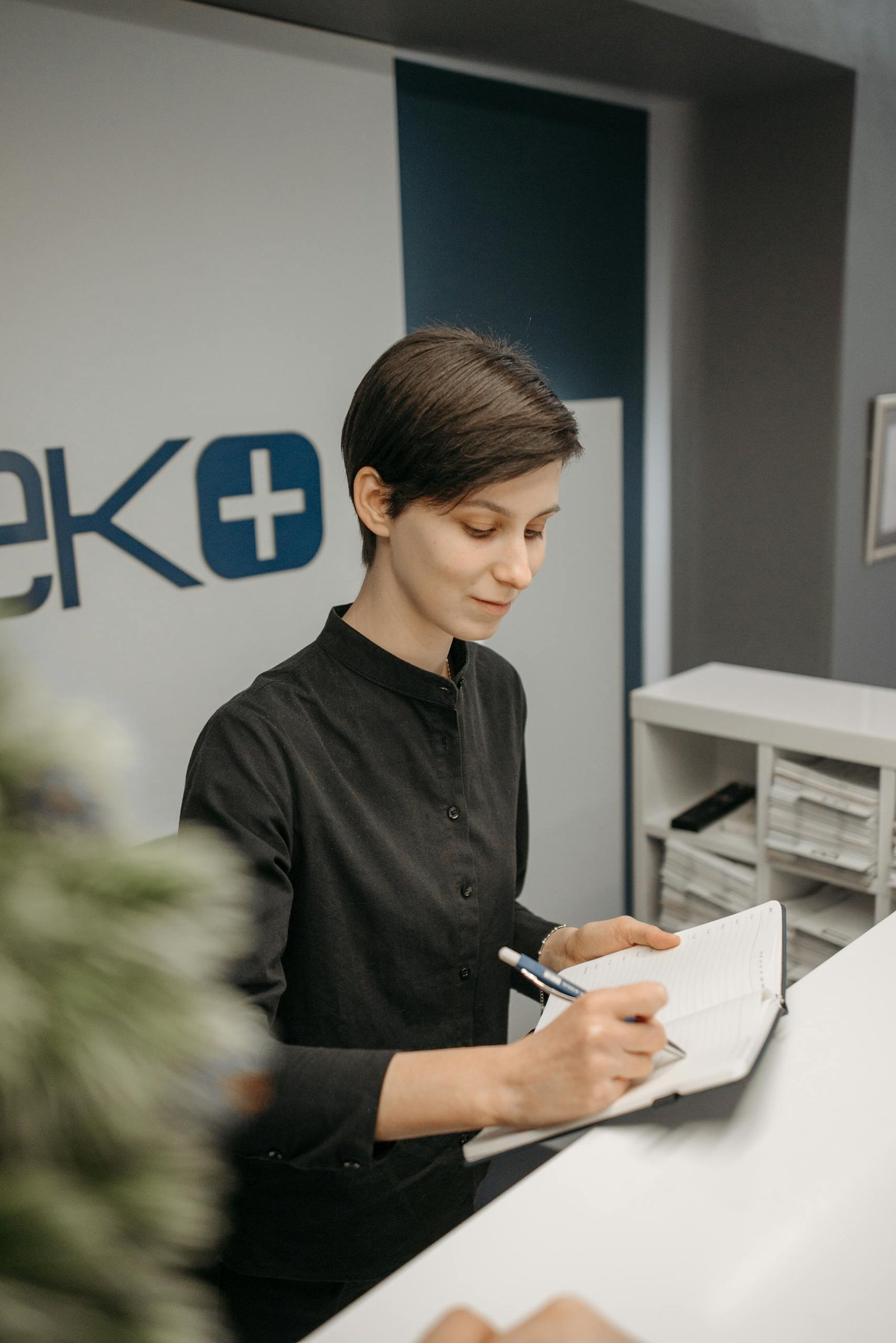 Female receptionist writing in a notebook at a clinic reception area, showcasing professionalism.