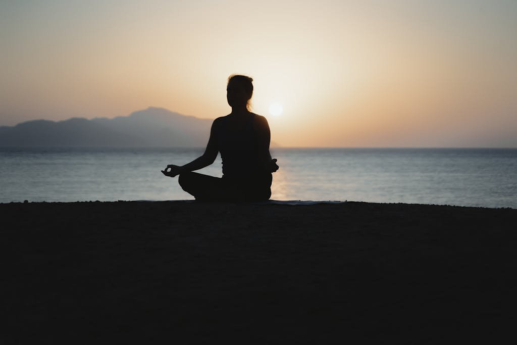A woman practices meditation with a tranquil ocean sunset backdrop, embodying peace and relaxation.